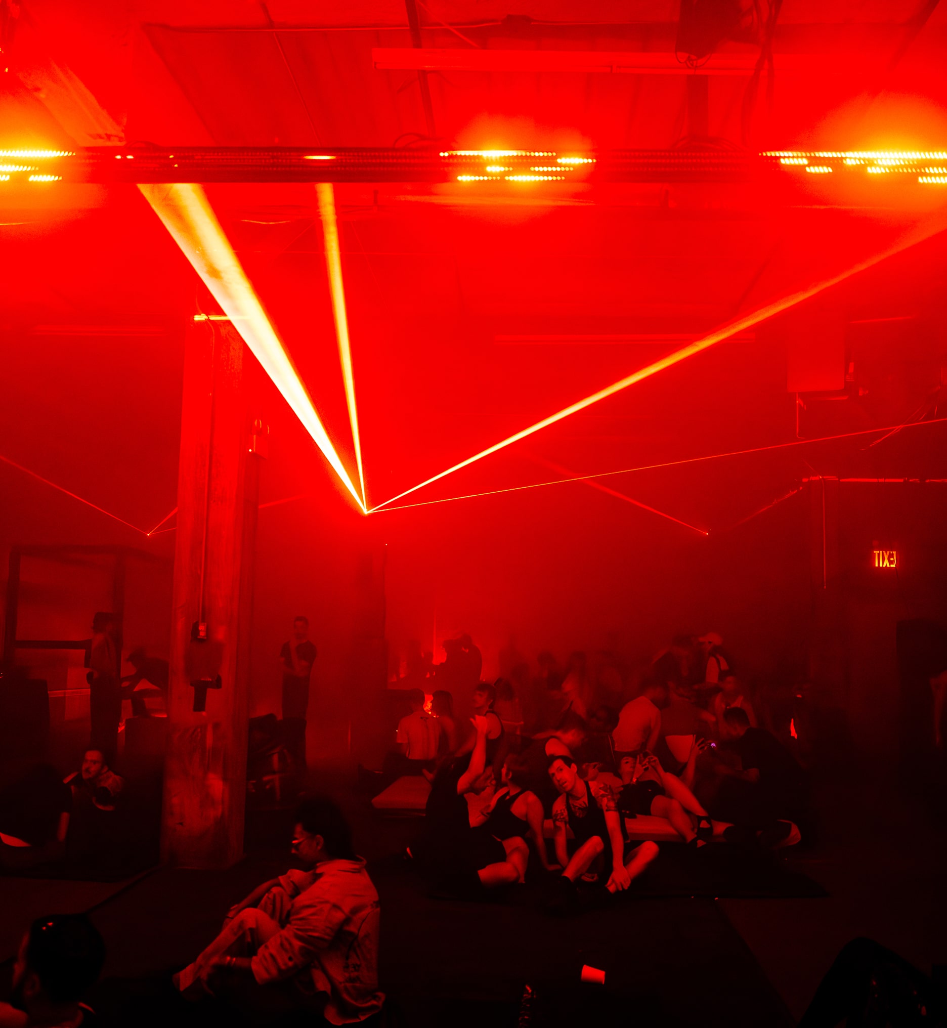 People lounging and talking under deep red light and lasers cutting through haze inside a warehouse dance space.